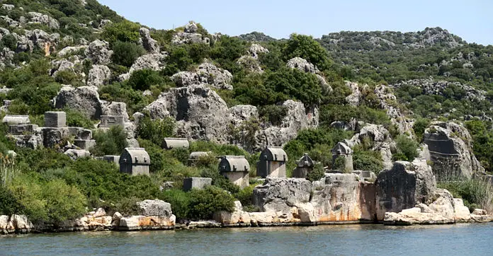 Lykische Steinsarkophage stehen an der Küste von Ücagiz am Meer auf einem kleinen grün bewachsenen Hügel.