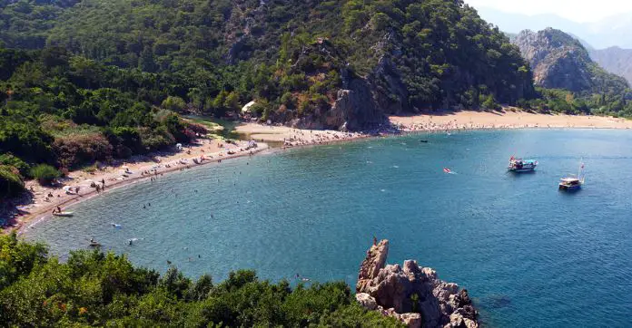 Panoramablick auf den Olympos Strand mit Badegästen an einer Flussmündung und Schiffen vor der Küste.