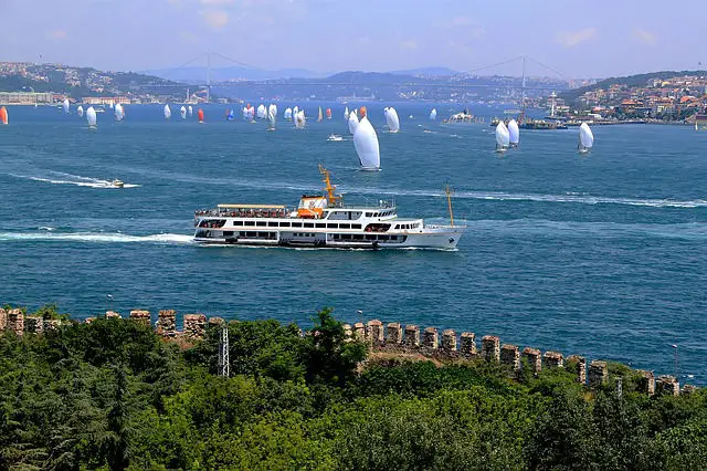 Viele kleine Segelschiffe am Bosporus in Istanbul. Im Vordergrund ist eine Stadtmauer und eine große Fähre zu sehen.