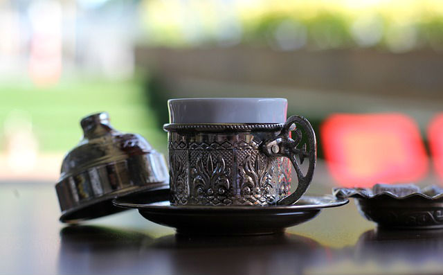 Tasse mit Türkischem Kaffee Metalluntertasse in die eine Porzellantasse mit türkischem Kaffee gestellt worden ist. Daneben liegt ein Deckel für die Tasse auf einem Tisch