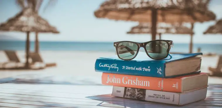 Drei Bücher in roter, blauer und weißer Farbe sind auf einem Holztisch aufeinandergestapelt. Auf den Büchern liegt eine Ray Ban Sonnenbrille. Dahinter ist ein Strand, Sonnenschirme, Sonnenliegen und das Meer zu sehen.