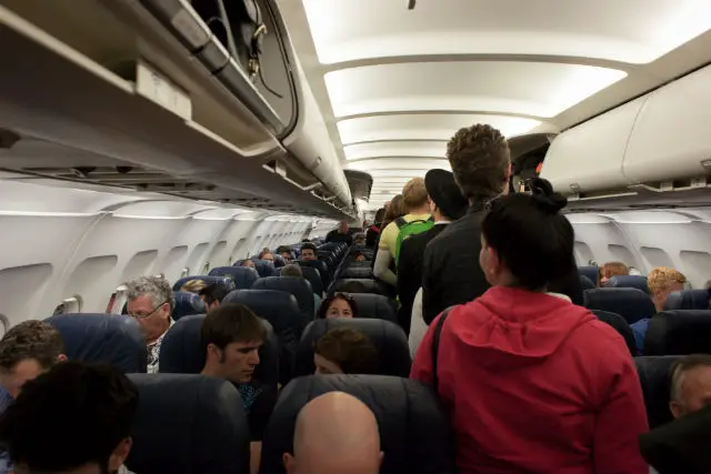 Das Innere einer Flugzeugkabine. Menschen steigen gerade in das Flugzeug ein und verstauen ihr Handgepäck.