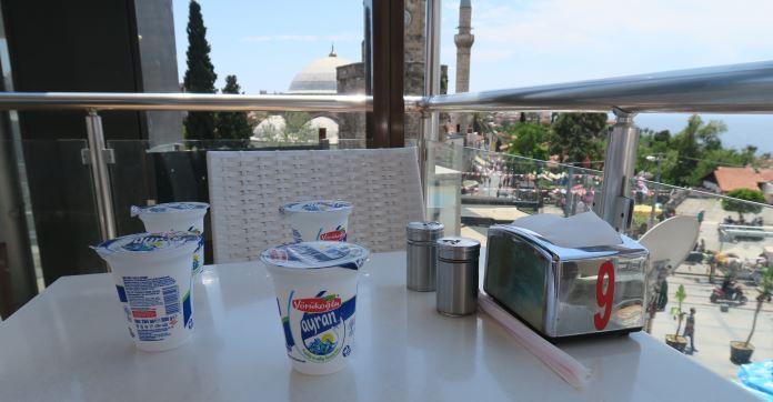 Vier Ayran Becher stehen auf einm Restauranttisch. Dahinter ist das Meer zu sehen und eine Moschee. 