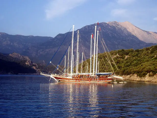 Ein Gulet ist ein türkisches Segelschiff. Es ist aus Holz gebaut. Die Masten sind weiß. Die Farbe der Segel ist nicht zu erkennen, weil sie eingerollt sind. Das Bild ist von einem anderen Schiff aufgenommen worden. Davor ist das blaue Wasser des Mittelmeeres zu sehen. Im Hintergrund befinden sich die hohen Berge des Taurusgebirges
