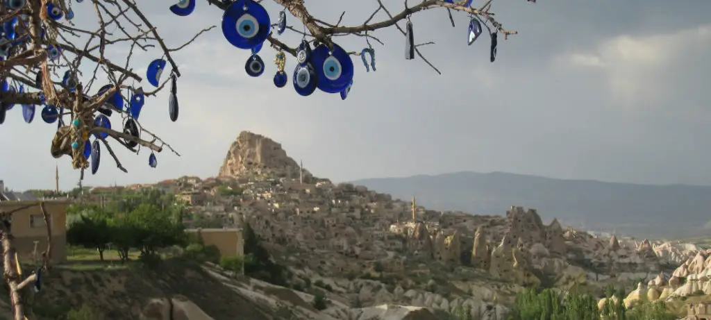 An einem Baum hängen türkische Augen (Nazar Amulett - klein augenförmig geformte Anhänger in blauer, weißer und schwarzer Farbe) an einem Baum. Im Hintergrund ist die sandfarbene Landschaft von Kappadokien mit ein paar grünen Bäumen vor der Festung Uchisar zu sehen.