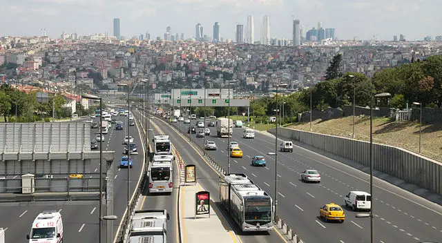 Aufgenommen von einer Straßenbrücke in Istanbul. Auf der Straße sind vier Busse der Metrobuslinie zu sehen. Der Blick geht der Straße entlang Richtung Innenstadt und zu den Hochhäusern der Sykline von Istanbul.