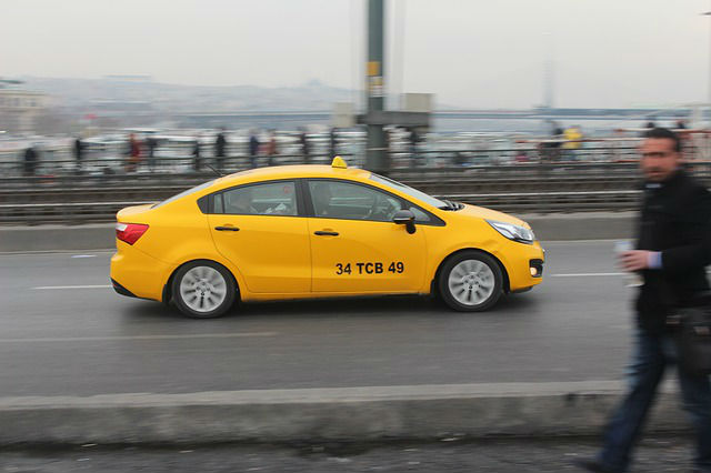 Ein während der Fahrt vom Straßenrand aus fotografiertes, gelbes Taxi in Istanbul. Das Taxi ist klar zu sehen. Die Umgebung ist wegen der Bewegung ein wenig verschwommen. 