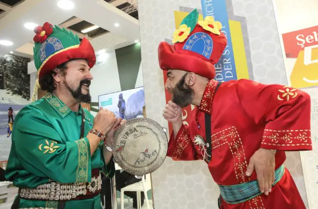 Zwei Türken in traditioneller Tracht am Stand von Bursa. Der eine trägt rote Kleidung und der andere grüne. Beide tragen einen Hut mit Blumen darauf. 