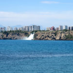 Düden Wasserfall aus zwei Kilometer Entfernung vom Yakamoz Strand in Antalya aufgenommen. Die hinter dem Wasserfall gebauten Hochhäuser wirken gleich hoch wie die Klippen