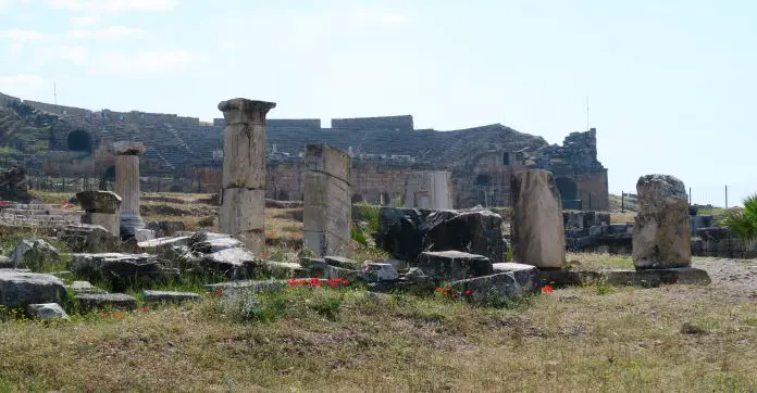 Amphitheater von Süden aus gesehen. Blick auf die Sitzreihen, vor dem Theater liegende Ruinen. Es blühen ein paar Rote Blumen auf der Wiese davor. 