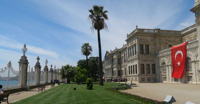 Die zum Bosporus ausgerichtete Seite des Dolmabahce Palast. Es ist eine Parkanlage mit kurz geschnittenem Rasen, Palmen, Sträuchern und dem weißen Palastgebäude zu sehen. Ein weißer Eisenzaun grenzt den Palast vom Meereswasser ab.