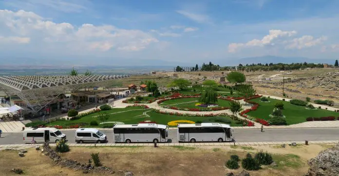 Blick von einem 10 Meter hohen Hügel auf den Eingangsbereich der Ruinen von Hierapolis. Auf einem Parkplatz stehen mehrere weiße Busse. Zwischen den alten Stadtmauern und den Bussen ist ein grüner Park mit vielen bunten Blumen zu sehen