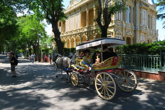 Kutsche auf Büyükada Kutsche mit zwei weißen Pferden fährt auf einer geteerten Straße an einem alten, weißen Holzhaus vorbei