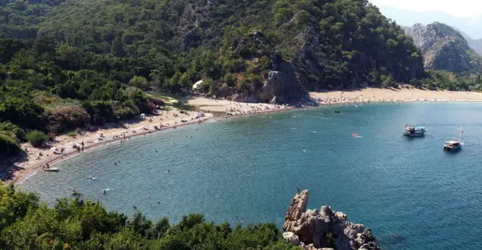 Blick auf den Olympos Beach in Cirali von einem nahe gelegenen Hügel. Es sind viele Badegäste am Strand zu sehen. Dahinter sind hohe Hügel mit Pinienwäldern zu sehen. 