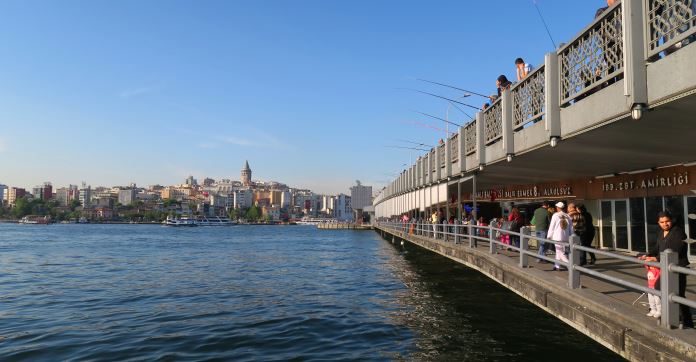 Fischer stehen auf der oberen Seite der Galatabrücke. Auf ihrer unteren Seite gehen Menschen über die Brücke. Auf der anderen Seite des Goldenen Horn ist der Galata Turm zu sehen. 