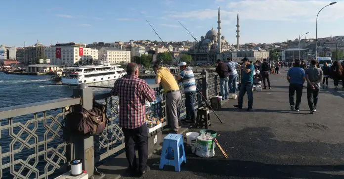 Fischer stehen mit ihren Angeleln auf der Galatabrücke. Am linken Bildrand ist eine Bosporusfähre zu sehen. Im Hintergrund steht eine Moschee. 