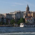 Galataturm rag mit seiner Spitze über die anderen Häuser Istanbuls heraus. Im Vordergrund sind mehrstöckige Häuser zu sehen und davor die Fährenanlegestelle Karaköy mit einer weißen Fähre.