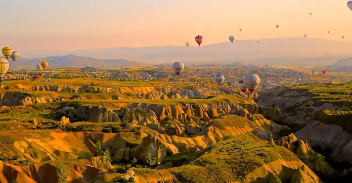Die zerklüftete Landschaft Kappadokiens mit ihren sandsteinfarbenen Schluchten, kärglich grünen Gras, Bergen und dutzenden Heißluftballonen in der Luft.