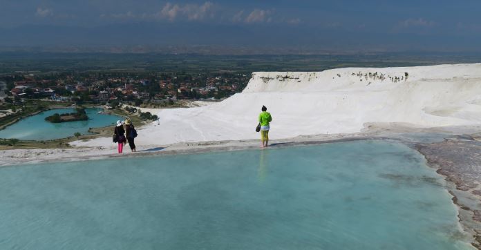 Blick vom oberen Rand der Wasserbecken auf die Kalkterrassen von Pamukkale, das türkisblaue Wasser darin, die schneeweißen Kalkablagerungen und den See darunter. 