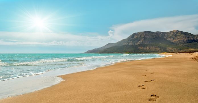 Fußspuren sind am Patara Sandstrand zu sehen. Der Blick geht Richtung Sonne. Die Brandung des tükisblauen Meereswassers ist zu sehen. Der Strand endet nach vielen Kilometern an einem Berg. 