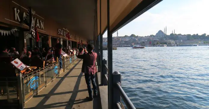 Restaurants unter der Galatabrücke Auf der linken Seite des Fußgängerwegs sind viele Restaurants mit Tischen und essenden Menschen zu sehen. Auf der rechten Seite schützt ein großes, weißes Sonnensegel entlang der ganzen Brücke vor der Sonne. Im Hintergrund ist die Süleymaniye Moschee zu sehen.