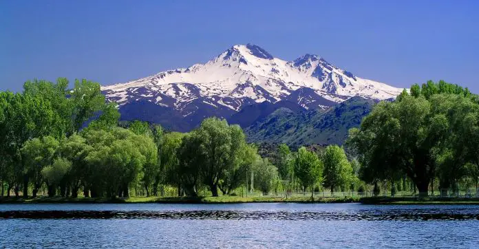 Blick vom Rand eines Sees auf einen dahinterliegenden Laubwald und den schneebedeckten Gipfel des Erciyes Dagi