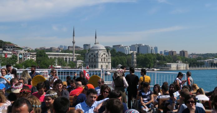 Dutzende Menschen sind auf dem Oberdeck einer Fähre zu sehen. Dahinter ist eine Moschee zu sehen.