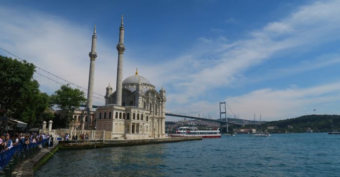 Die Ortaköy Moschee am Bosporusufer, neben der Bosporusbrücke. Fischer stehen am Ufer mit ihren Angelrufen. Die Sonne scheint.