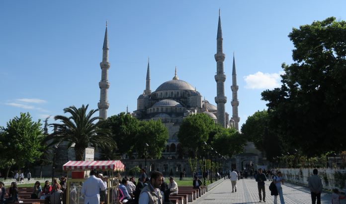 Der Platz vor der Moschee mit einem Simitverkäufer und seinem Stand, Touristen und den mit grauen Steinplatten ausgelegten Weg zur Blauen Moschee. Sie ist im Hintergrund zu sehen.