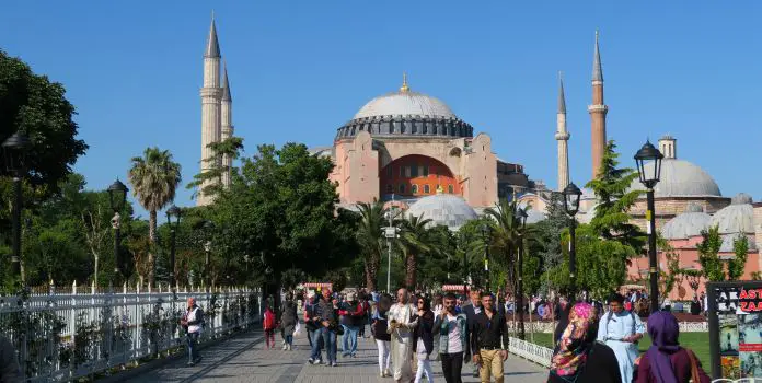 Der geplfasterte Weg zur Hagia Sophia durch einen Park. Es gehen viele Menschen auf dem Weg. Im Park wachsen Pinien und Palmen. 