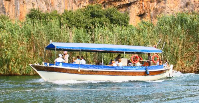 Ein blau-weiß-braunes Boot fährt mit einigen Touristen am Dalyan Fluss zum Strand.