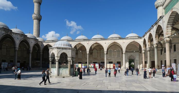 Der Innenhof der Blauen Moschee in Istanbul: Mit Säulengängen rund um einen zentralen Brunnen. Die Farbe der Steine ist ein leichtes Sandgrau. Der Boden ist mit Marmorplatten ausgelegt. Es sind einige Besucher der Moschee am Platz zu sehen. 