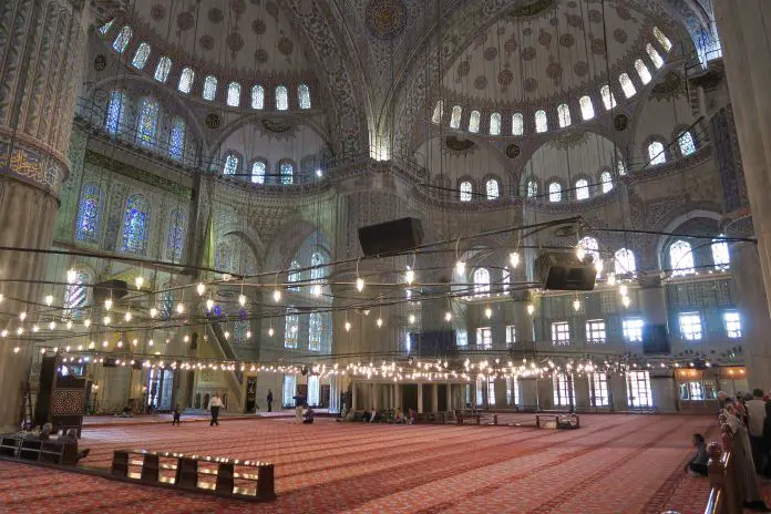Der rote Teppichboden im Gebetsbereich für Männer in der Blauen Moschee. Er ist unter der großen Kuppel. Es sind ein paar betende Männer zu sehen.