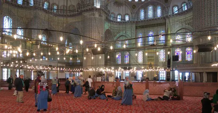 Ein Luster hängt an vielen Ketten von der 40 Meter hohen Kuppel der Blauen Moschee. Er hängt nur 2 Meter über den Köpfen der vielen Besucher in blauen Kopftüchern und Röcken. 