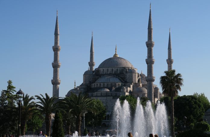 Das meterhoch hinaufspritzende Wasser des Springbrunnens vor der Blauen Moschee. Im Park sind Palmen und viele andere Bäume zu sehen, dahinter ist die Moschee. 