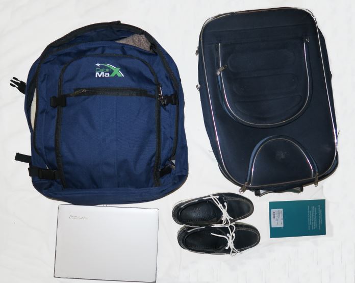 Ein Cabin Max Metz Rucksack, ein 15 Zoll Notebook, Sebago Bootsschuhe, ein Buch und ein normaler Koffer liegen auf einem Bett.