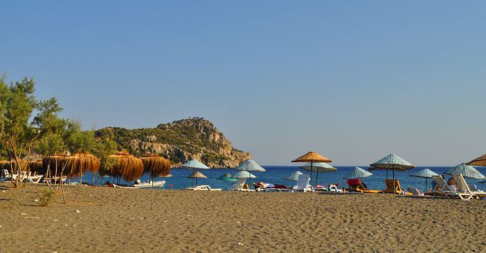 Der Strand von Datca. Reisende liegen auf Sonnenliegen, unter Sonnenschirmen. Der Blick geht Richtung Meer.