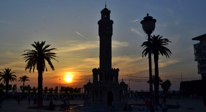 Der Uhrtrum von Izmir bei Sonnenuntergang. Daneben stehen Palmen und das Meer ist im Hintergrund zu sehen.