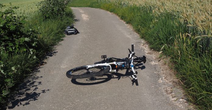 Ein Rad ist auf einem schmalen Feldweg umgefallen. Es liegt Mitten am Weg. Daneben sind Felder. 