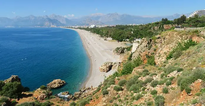 Der kilometerlange Konyaalti Strand im Antalya im April. Am Strand baden die ersten Badegäste, die Sonne scheint, das Gras, Bäume und die Palmen sind grün. 