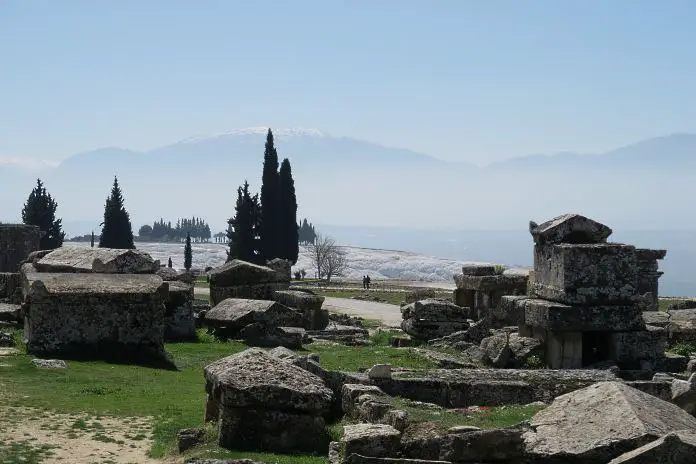 Römische Steinsarkophage stehen im Gras, im Hintergrund sie die schneeweißen Kalkterrassen von Pamukkale zu sehen, Pinien und die schneebedeckten Berge rund um Denizli im Hintergrund. 