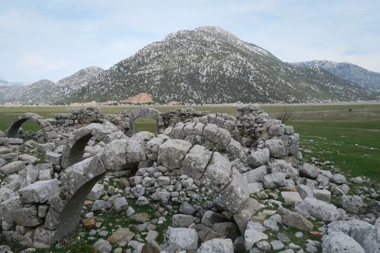 Die Ruinen der Karawanserei Tol Han bestehen aus alten Steinbögen und Mauern. Der Blick reicht auf ein mit Gras bewachsenes Hochplateau und auf schneebedeckte Berge. 