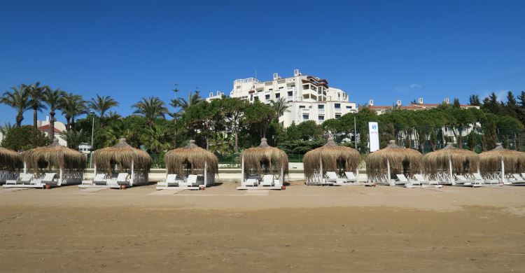 Ein weißes Hotel steht in einem Park mit Palmen. Am menschenleeren Sandstrand sind Liegestühle in kleinen Strandhütten zu sehen.