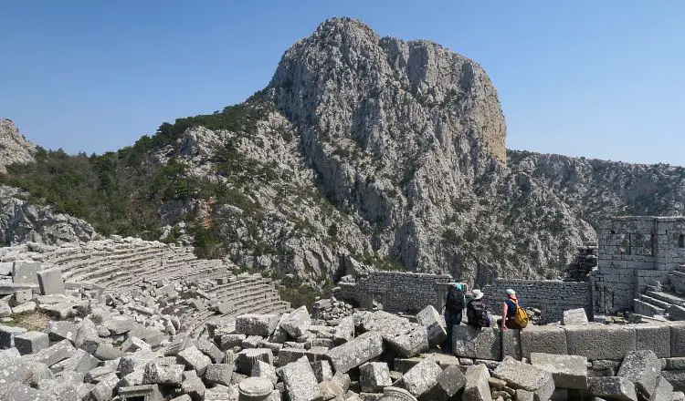 Das Theater steht auf einem Berggipfel im Taurusgebirge. Zu sehen sind die 5.000 Sitzplätze mit verstreut liegenden Steinen und Reisenden die auf dem obersten Rang des Theaters sitzen. Sie wirken klein. 