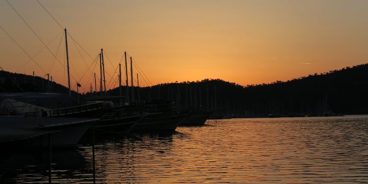 Die rötliche Sonne geht am Yachthafen von Fethiye hinter einem Hügel unter. Davor sind Schiffe und das Meer zu sehen.