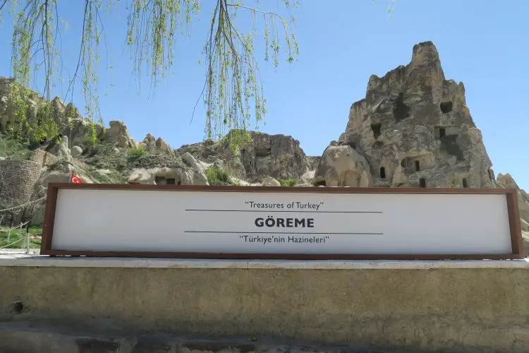 Am weißen Eingangsschild steht "Göreme Nationalpark". Dahinter sind Bäume und die Felsenkirchen im Park zu sehen.