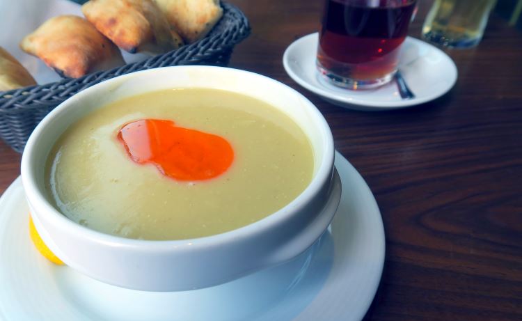 Eine weiße Schüssel mit der türkischen Suppe Mercimek Çorbası. Sie besteht aus Linsen. Daneben liegt Brot und es steht ein Glas mit türkischen Tee am Tisch.