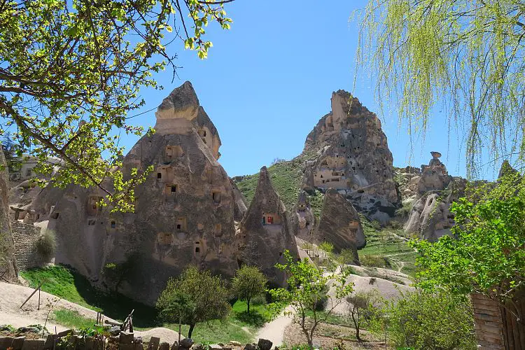 Du siehst die in den Fels gehauenen Festungsteile der Festung Uchisar und den grünen Garten mit Bäumen im Peri Cafe Cave Man. 