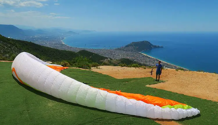 Ein Mann hält einen Paragliding-Schirm in seiner Hand der auf dem Boden liegt. Der Blick geht von der Spitze eines Berges bis weit hinaus auf das Meer und die Innenstadt von Alanya.