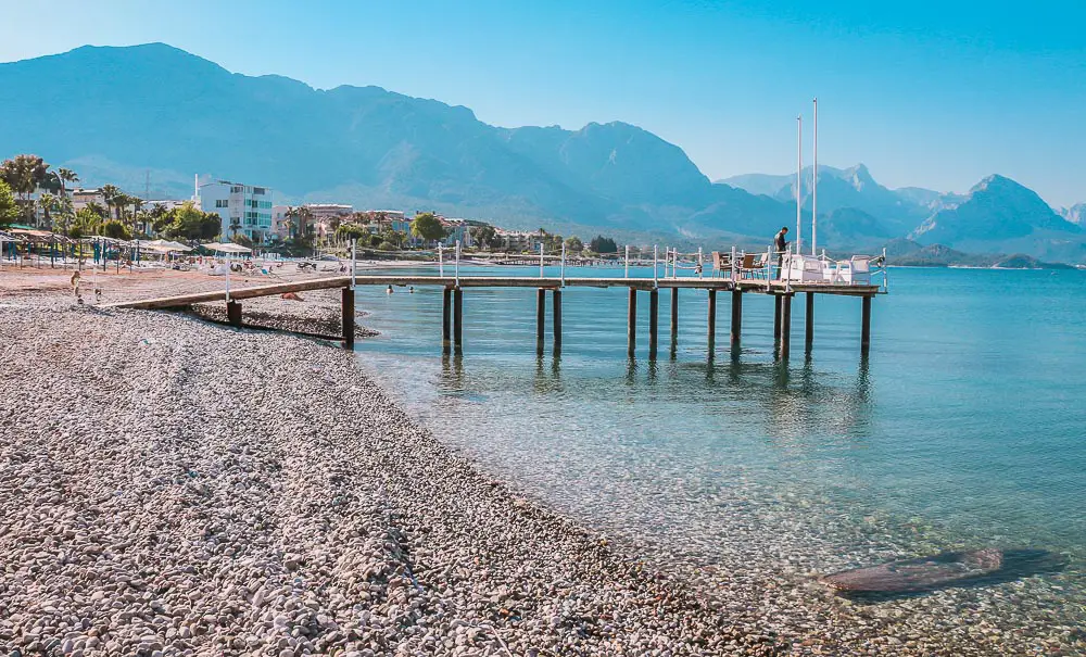 Kieselstrand mit einem Pier und dem Blick auf das Taurusgebirge mit der Innenstadt von Kemer